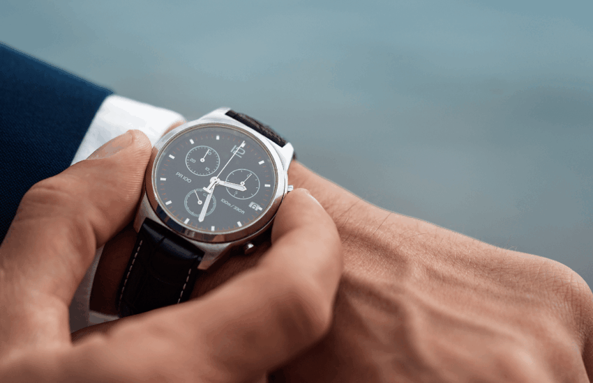 Close-up of a person adjusting a black leather–strap wristwatch on their left wrist, with a blurred blue background.
