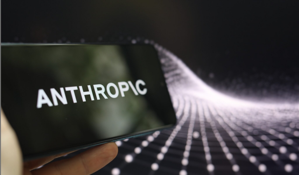 image - Trade Info Close-up of a smartphone screen displaying the word ANTHROPIC, held at a shallow angle against a dark, dotted background.