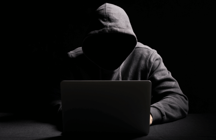 Hooded person with face obscured, sitting at a laptop in a dark setting.