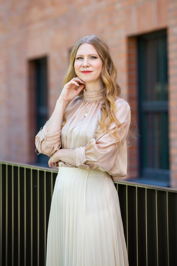 Olívia_Lacenova_photo - Trade Info - Forex.sk Woman with blonde hair in a beige blouse and cream pleated skirt leaning on a railing beside a brick building.