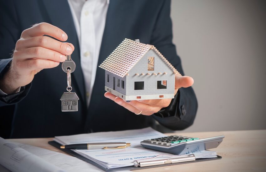 Businessperson holding a miniature house and a key over a desk with documents and a calculator.