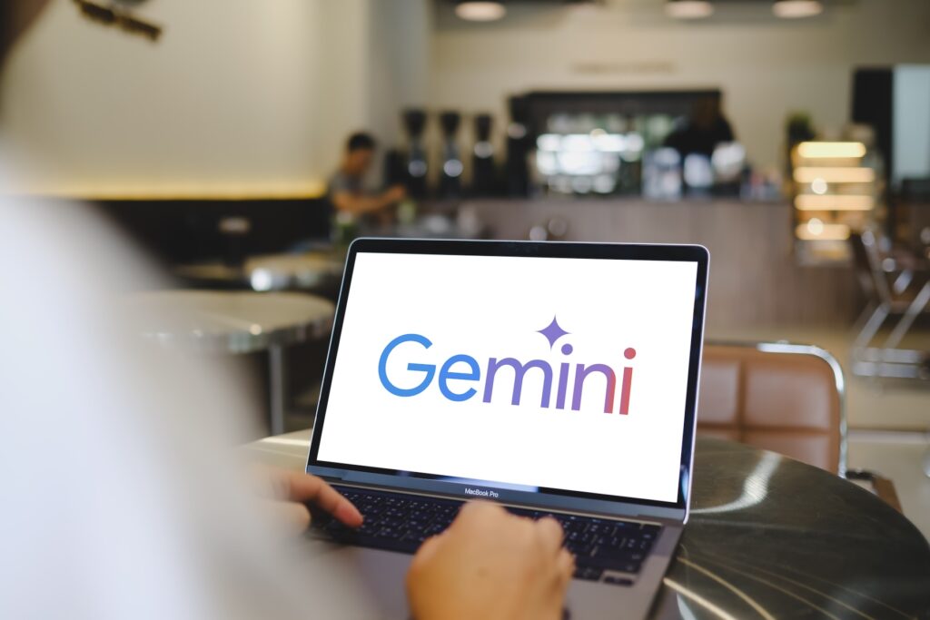 Gemini - Trade Info Person typing on a MacBook Pro with a bright Gemini logo on screen in a cafe setting.