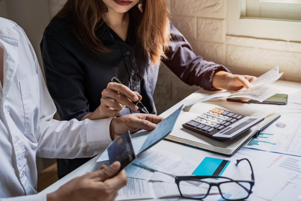 Two professionals review financial documents at a desk, using a calculator and pointing at papers.