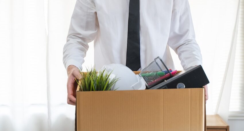 Person in a white shirt and black tie packs a cardboard box with office items (plant, hard hat, binder, organizers) for a move or relocation.